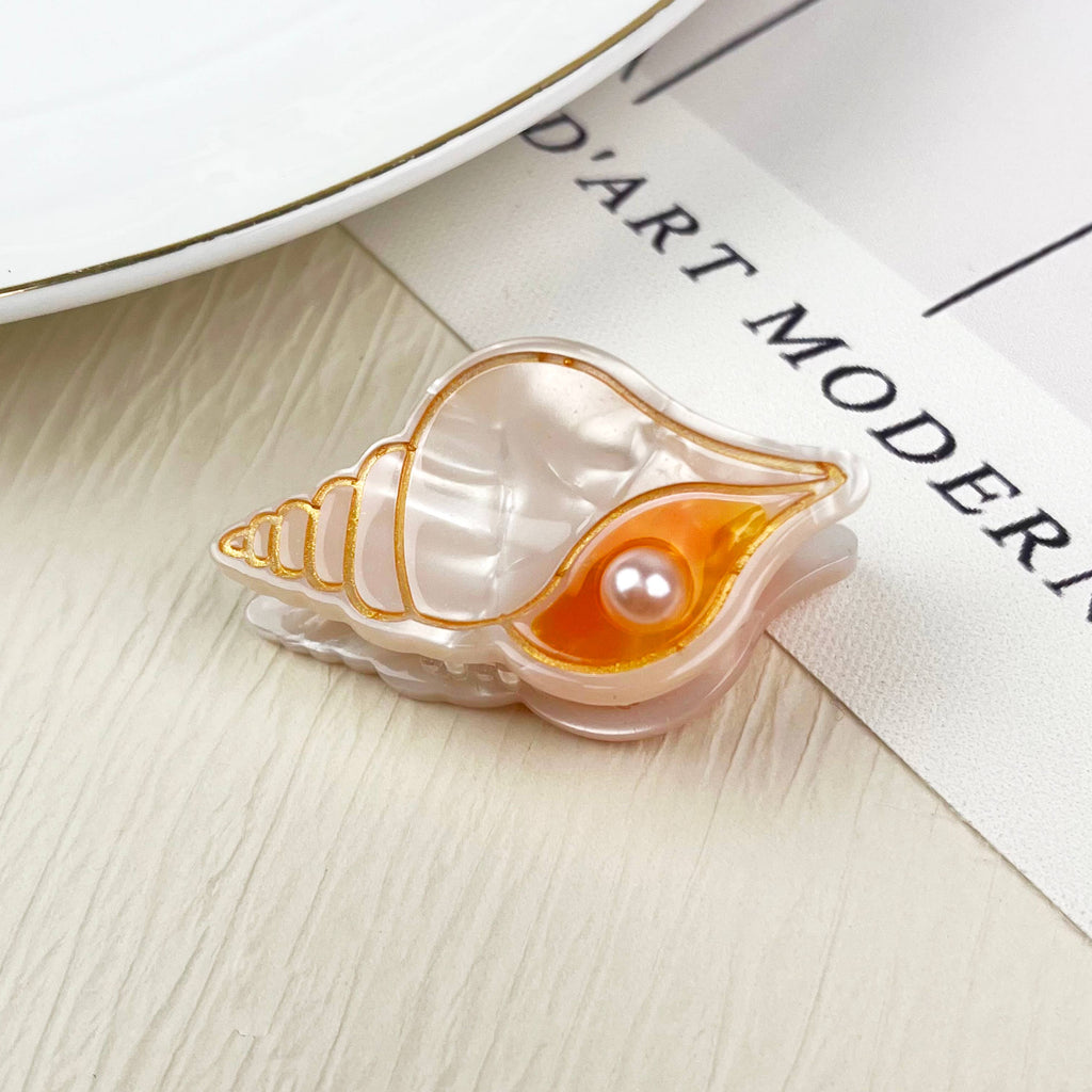 Chic mini hair clip designed as a translucent seashell with gold trim, an orange detail, and a pearl embellishment.