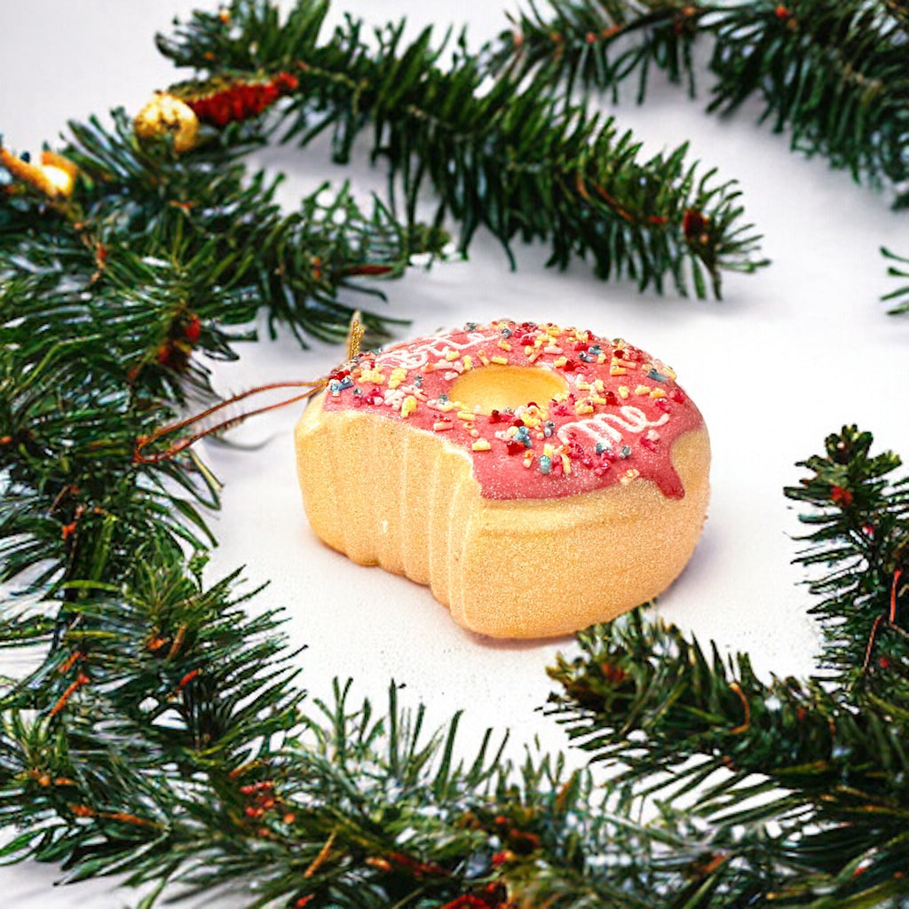 Playful holiday ornament shaped like a pink donut with "Bite Me" text and colorful sprinkles, hanging from a Christmas tree branch.