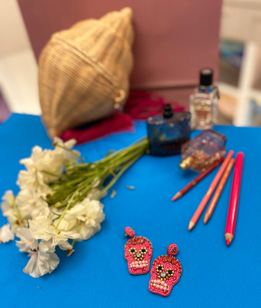 Vivid sugar skull earrings take center stage in this artistic flat-lay, surrounded by a vibrant mix of florals, warm-toned pencils, and luxe perfume bottles—perfect for showcasing bold, playful style in a creative setting.